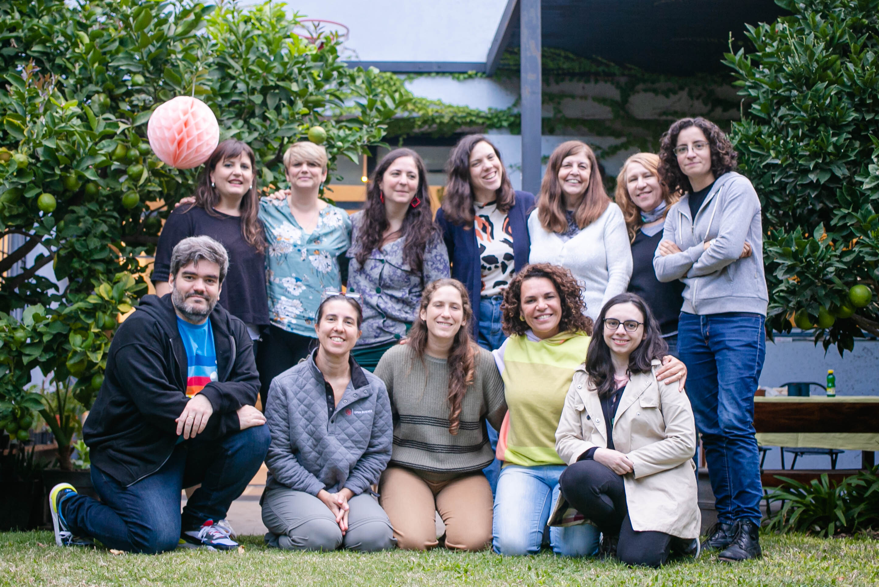 The image shows 12 people of different ages, informally dressed, in a natural garden, smiling and happy.