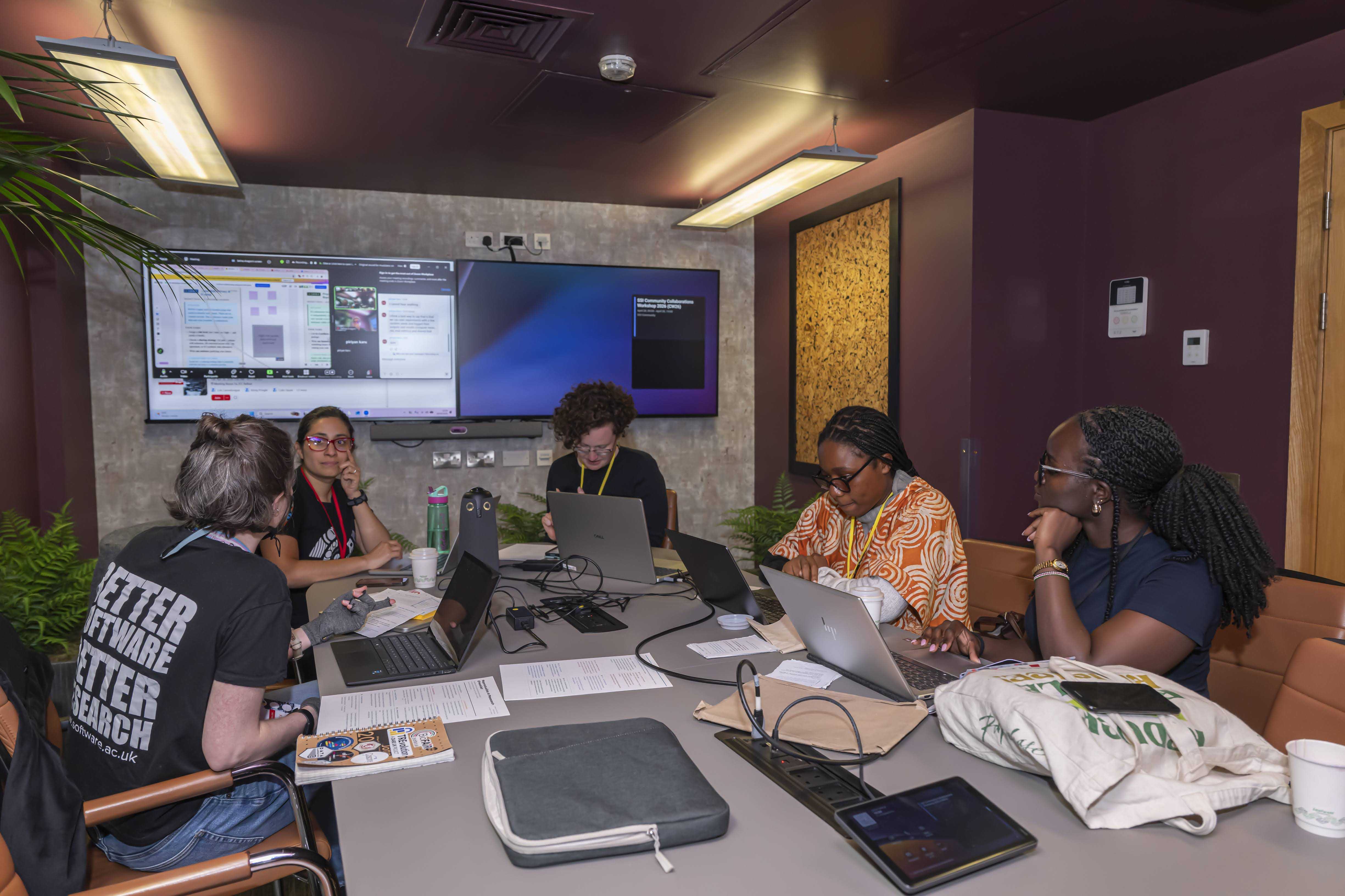 Five people co-working in a bordeaux and grey room around a table with computers, papers, bags, outlets and other equipment. Two screens on the back wall.
