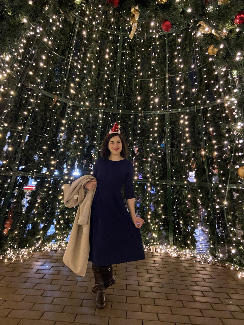 At the front of the image: Gracielle is wearing a long blue dress and a Christmas hat, holding her coat, very smily, posing inside a big Christmas tree