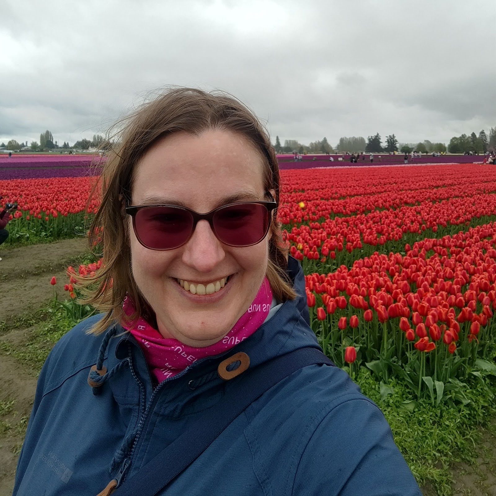 At the front of the image: Laura is very smily woman, wearing sunglasses and a raincoat at the Skagit Valley Tulip Festival in April 2025. Red tulip fields can be seen at the background