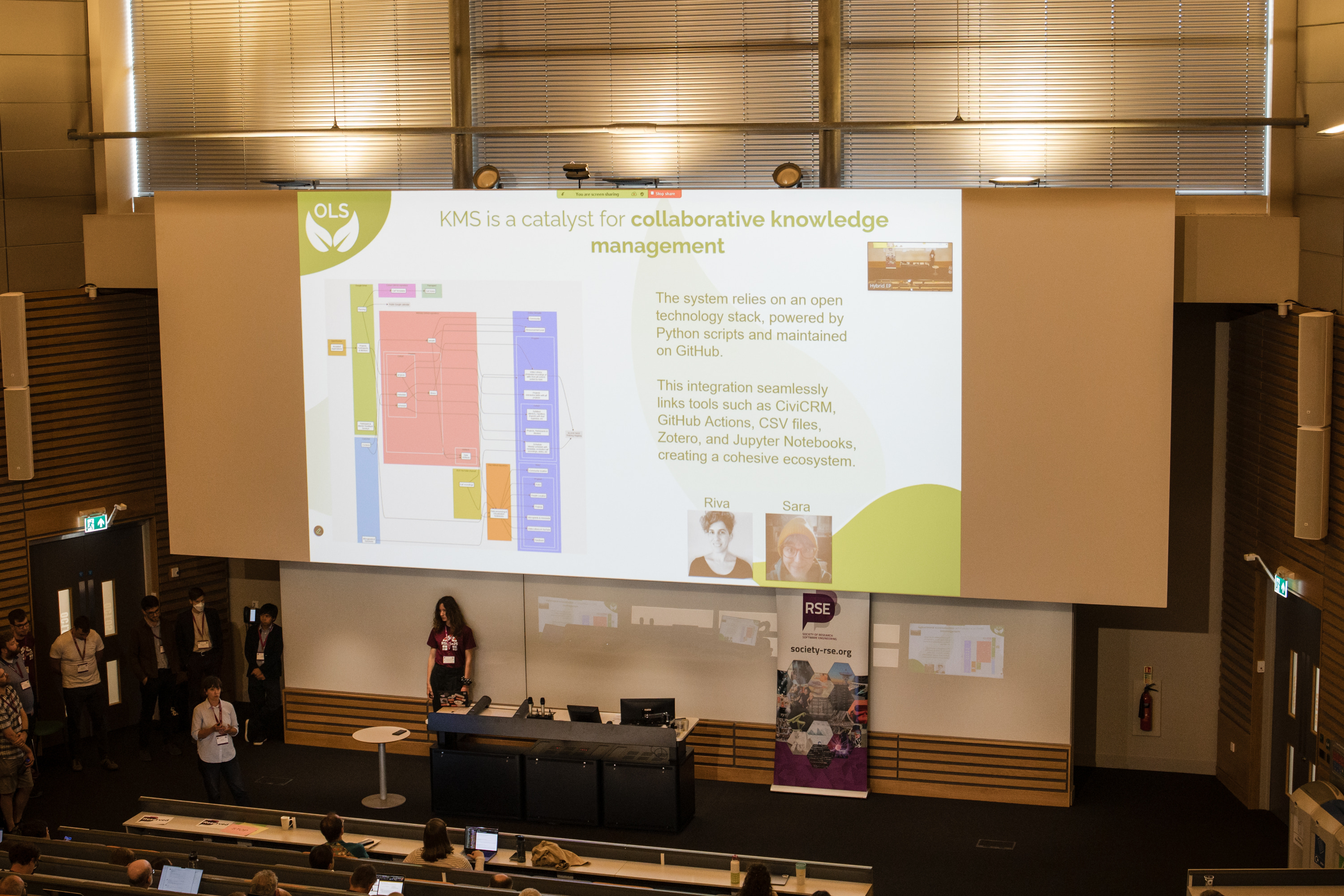 Image shows a shot from the back of the room, where Sara is delivering her talk. She appears very small in front of a very big screen showing the KMS graphic