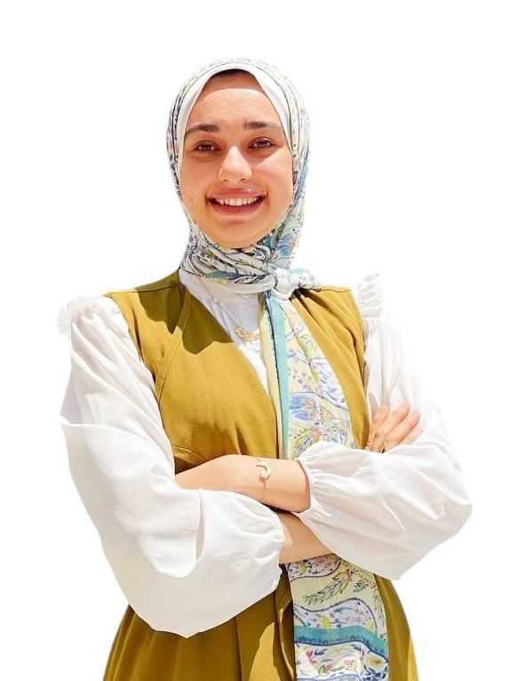 Image shows Doaa, an Egyptian woman, wearing a white long-sleeved blouse, a sleeveless mustard dress and a floral hijab. She has her hands cross in front of her, with a broad smile on her face.