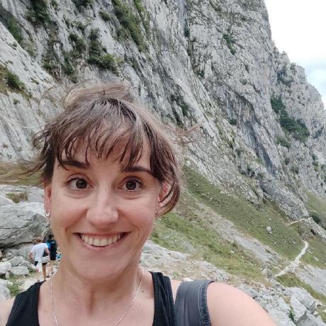 Image shows Sara Villa, a woman from Palencia, Spain. She has brown bangs, and is wearing a black tank top, with the strap of a black backpack slung over one shoulder. Sara is posing infront of some mountains in Picos de Europa National Park, with wide eyes and a broad smile!