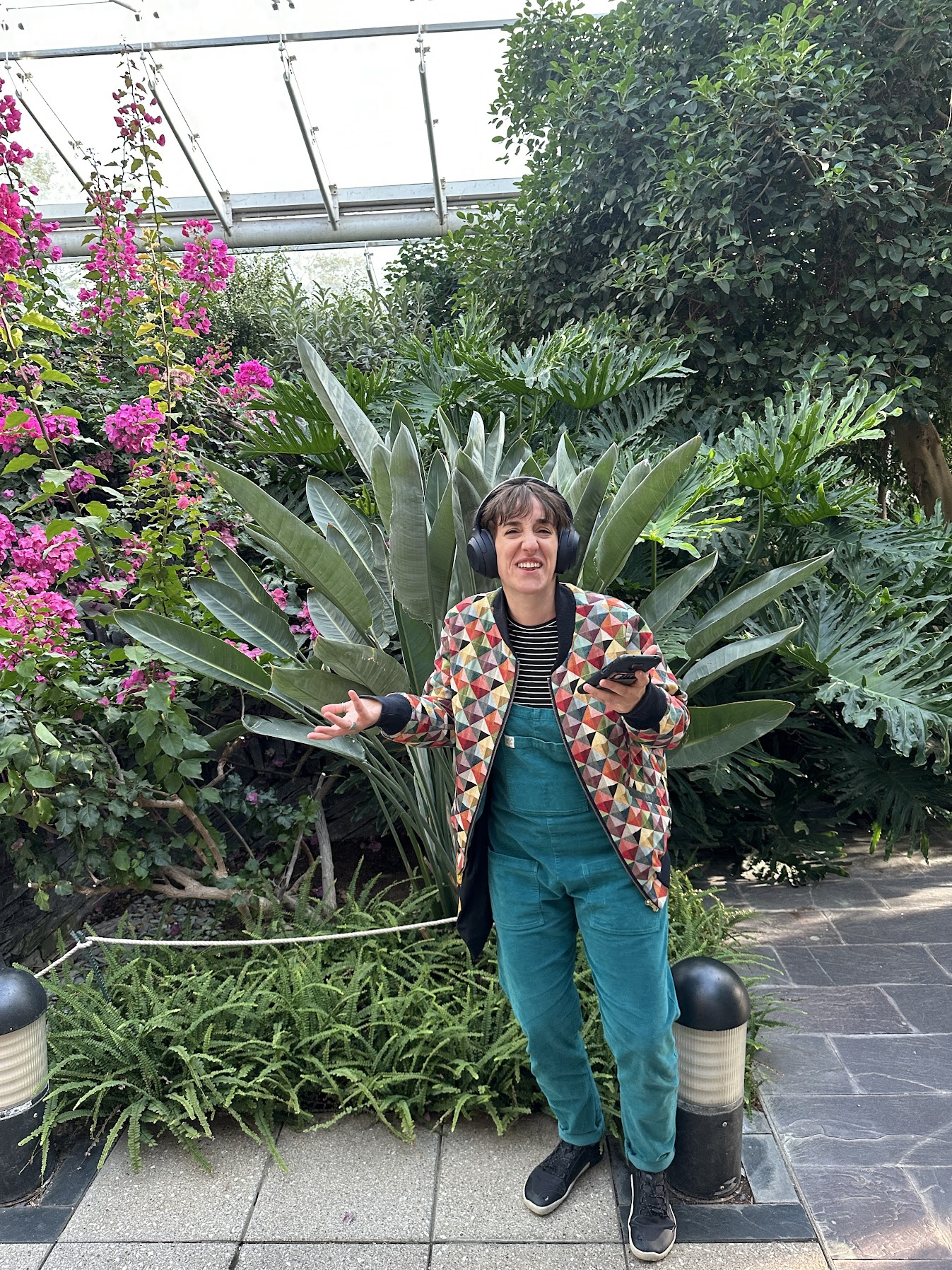 Sara is wearing headphones and colorful clothing, standing among tropical plants and pink flowers in a greenhouse, holding a smartphone.